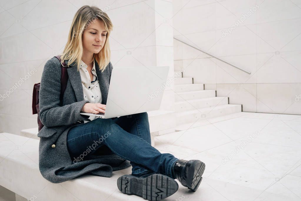 young attractive blonde female student using laptop while sitting on the stone bench. copy space area for your text message or content