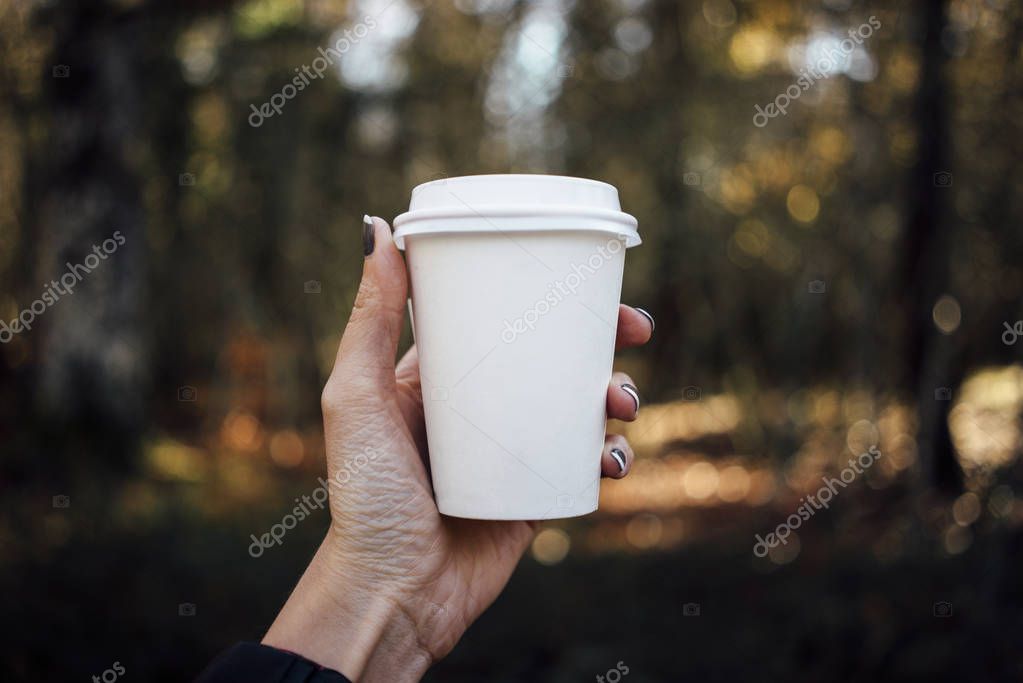 Female hand holding a blank paper cup with plastic cap on forest background