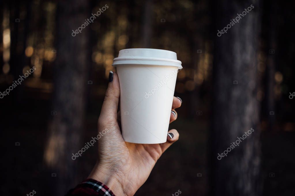 Female hand holding a blank paper cup with plastic cap on forest background