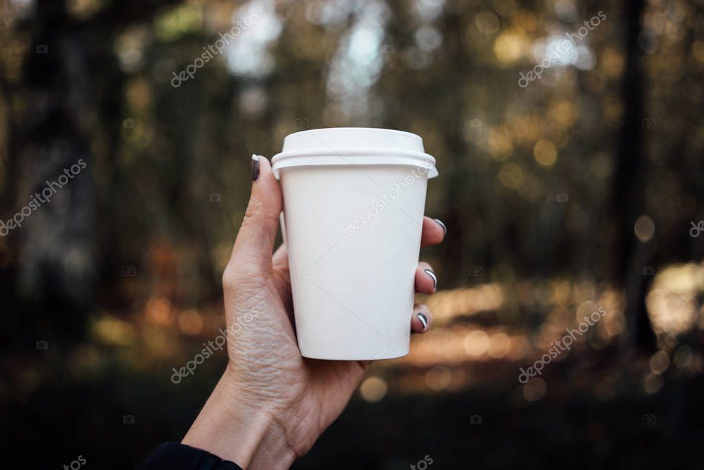 Female hand holding a blank paper cup with plastic cap on forest background