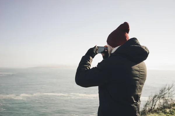 young man wearing a jacket and hat doing a photo while standing on a cliff by the ocean