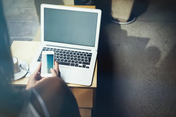 Cropped image of young woman using smartphone while sitting at table with laptop