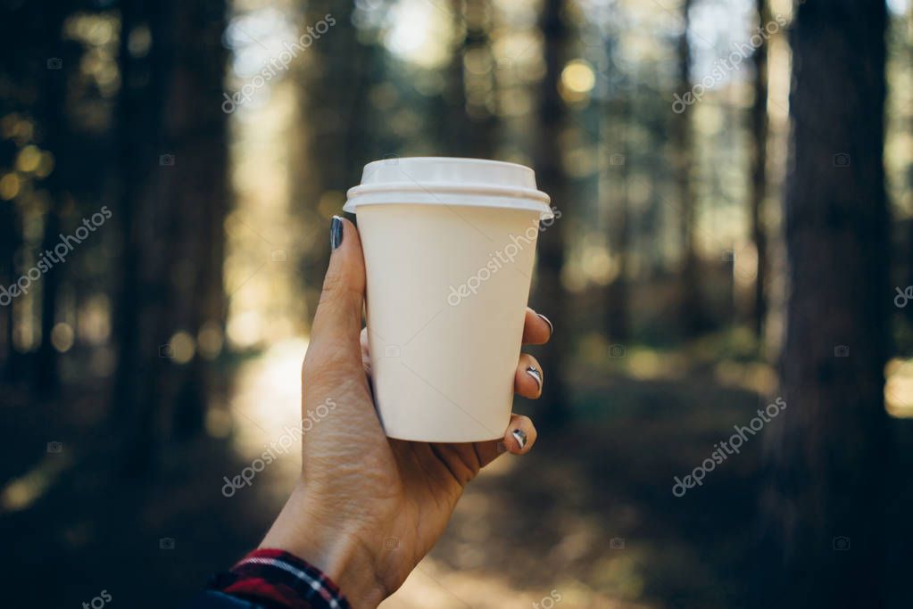 Female hand holding a blank paper cup with plastic cap on forest background