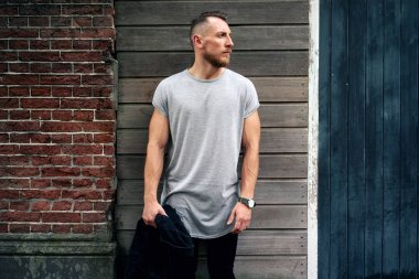 yakışıklı kaslı adam boş gri t-shirt giyen bir kenara tuğla ve ahşap duvar bir sokak yanında dururken arıyor