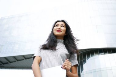 beautiful Asian woman with laptop posing gainst modern scyscraper clipart