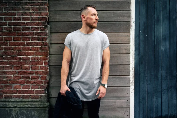 yakışıklı kaslı adam boş gri t-shirt giyen bir kenara tuğla ve ahşap duvar bir sokak yanında dururken arıyor