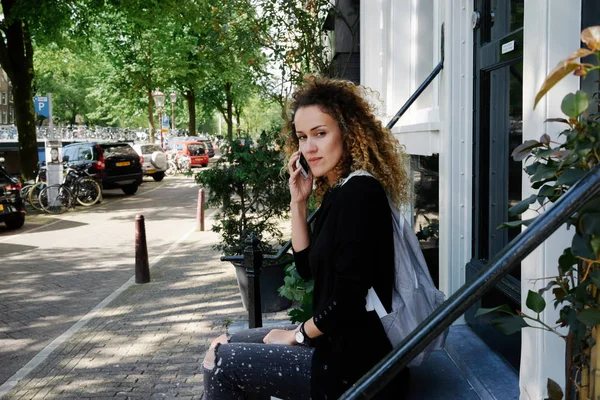居心地の良い都市に坐っている間通りスマート フォンで話して巻き毛を持つ魅力的な若い女性 — ストック写真