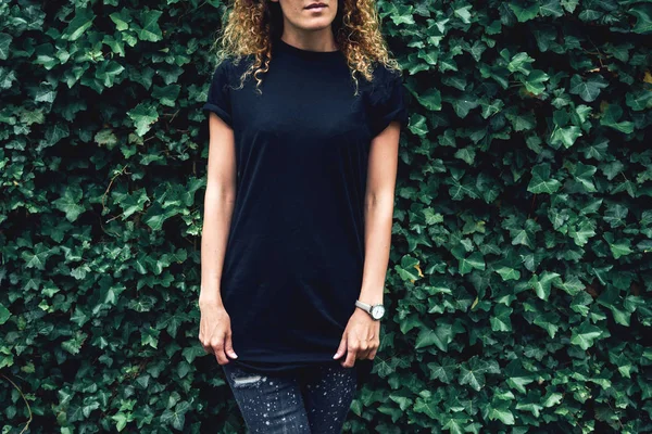 Cropped image of young hipster woman wearing blank black t-shirt and black jeans standing on a plants background