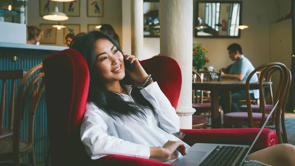 カフェでノート パソコンで作業しながらスマート フォンで話している若い美しいアジアの女性 — ストック写真
