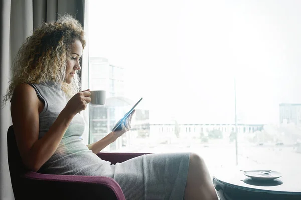 beautiful thoughtful woman with a cup of coffee in the hand wearing long gray dress sitting near big window of a modern cafe and holding a digital tablet