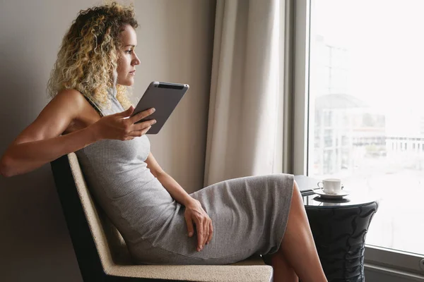 一杯のコーヒーとホテルのロビーで座りながらデジタル タブレットを使用して巻き毛を持つ魅力的な女性実業家 — ストック写真