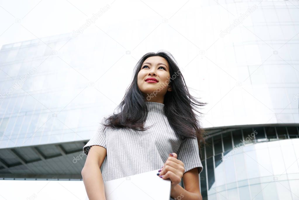beautiful Asian woman with laptop posing gainst modern scyscraper