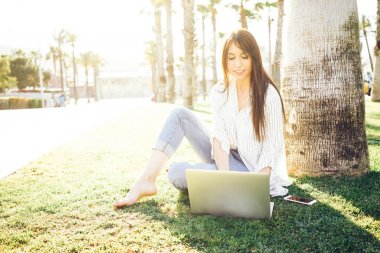 Çizgili bir gömlek genç kadın parkta otururken bir dizüstü bilgisayarda web sörf yapıyor 