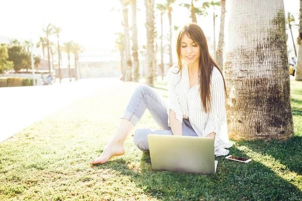 ストライプのシャツを着た若い女性が公園に座りながらノート パソコンで Web をサーフィンします — ストック写真