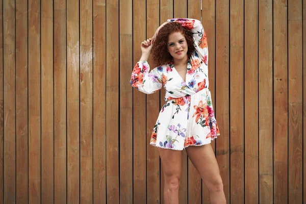 A curly red haired woman with freckled face wearing short colored dress posing on a street beside wooden fence on a sunny day