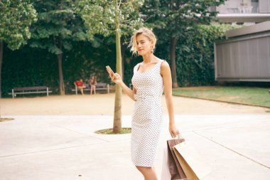 Beyaz yazlık elbise siyah lekeli ile duran smartphone ile park boş kağıt torbalar ile giyiyor güzel beyaz kadın