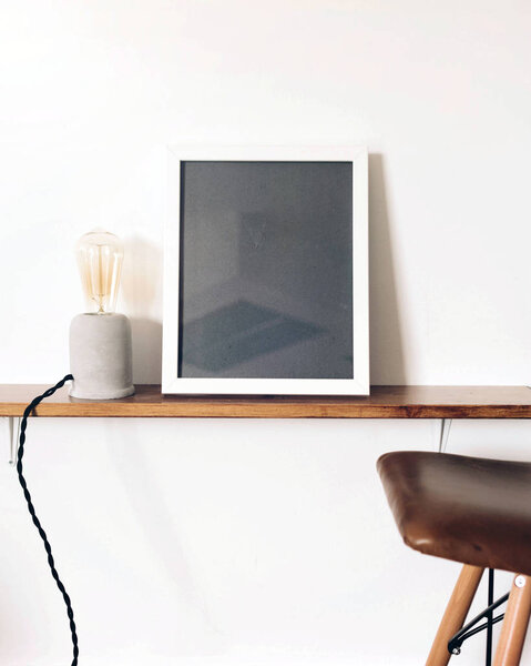 White room with empty white frame standing on the massive wooden shelf. Retro lamp and chair with an vintage leather seat. Decoration of the interior of the studio