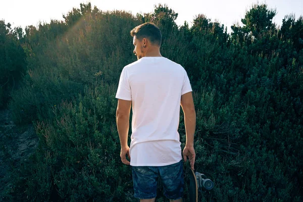 Young male wearing a blank white t-shirt standing with his back to camera. A man with a scateboard is looking aside on a plant wall background