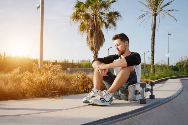 Young bearded man in a street wear is sitting with the skateboard in the urban zone. A skater is having rest in the urban zone on a sunset