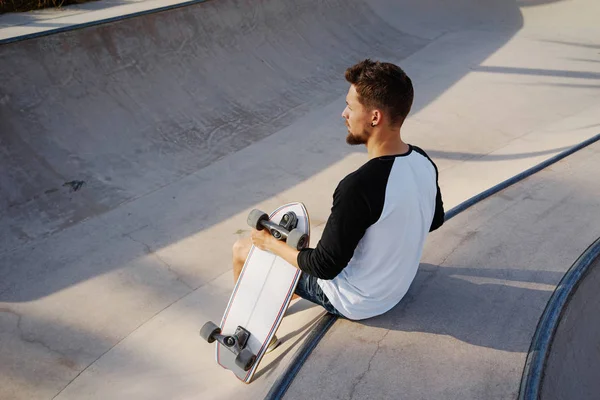 Young bearded man in a street wear is sitting with the skateboard in the urban zone. A skater is having rest in the urban zone on a sunset