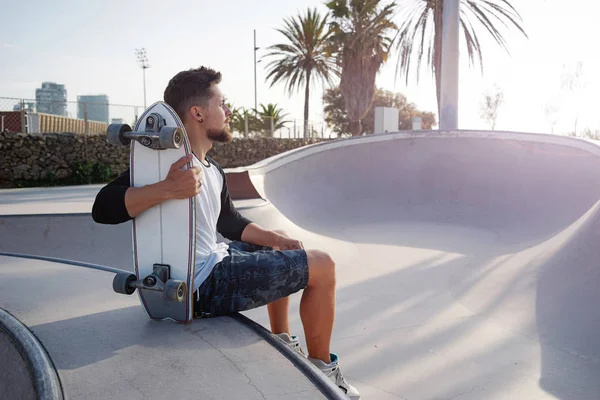 Young bearded man in a street wear is sitting with the skateboard in the urban zone. A skater is having rest in the urban zone on a sunset