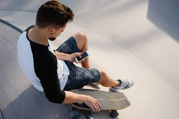 若い男は スケート ゾーンに座りながら携帯電話を保持しています 夕日を背景に座りながらスマート フォンで Web をサーフィンします — ストック写真