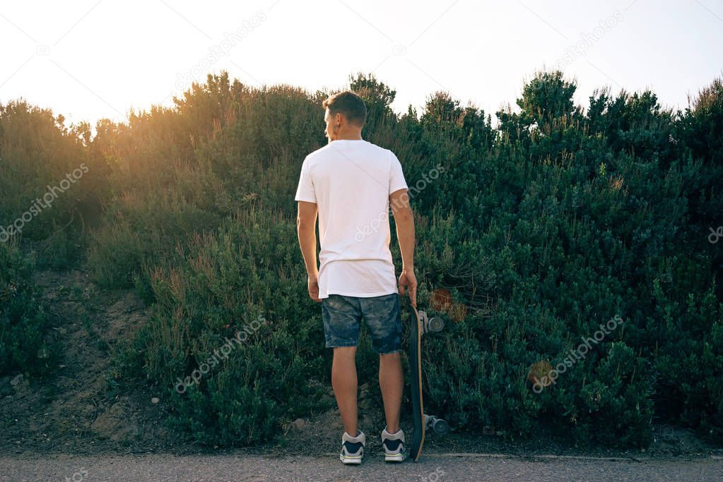 Young male wearing a blank white t-shirt standing with his back to camera. A man with a scateboard is looking aside on a plant wall background