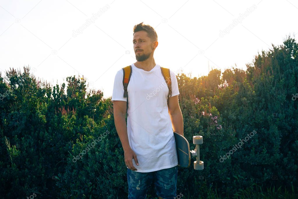 Young man with a skateboard in a blank white t-shirt standing in a bushy area