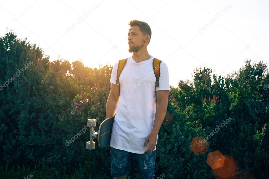 Young man with a skateboard in a blank white t-shirt standing in a bushy area