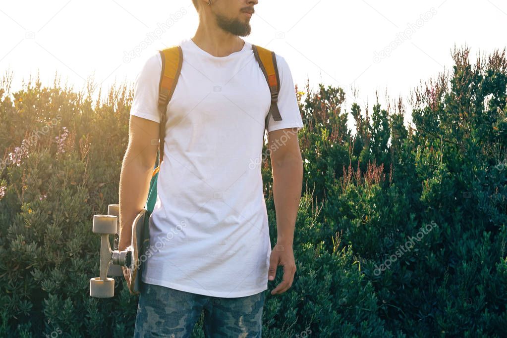 Young man with a skateboard in a blank white t-shirt is standing in a park