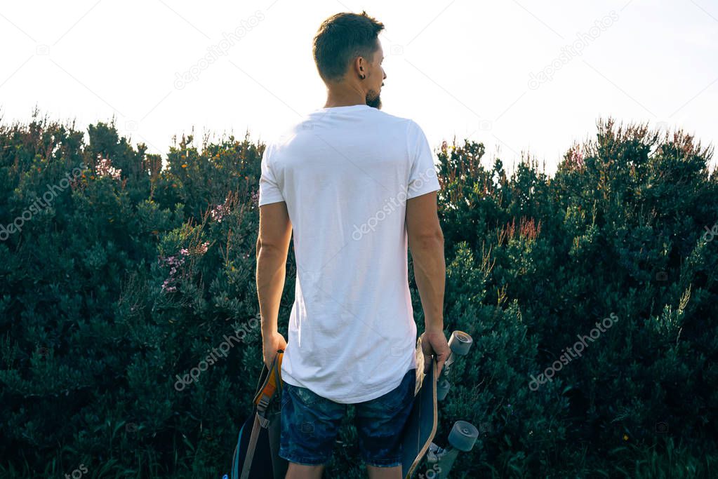 Young male with a scateboard in a blank white t-shirt is standing with his back to the camera. A man with a backpack is looking aside on a plant background.