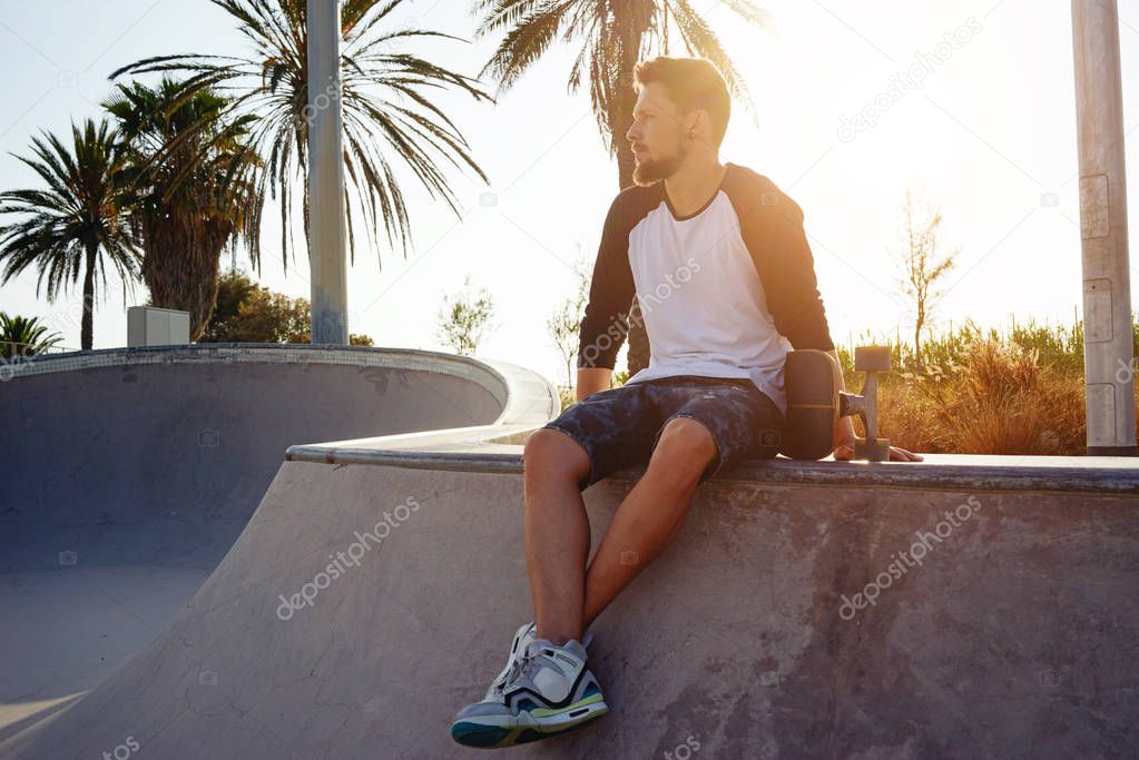 Young bearded male in a street wear is sitting with the skateboard in the skate zone 