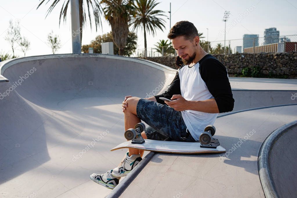 Young man is holding a mobile phone while sitting in a skate zone. A guy is surfing the web by the smartphone while sitting on a sunset background