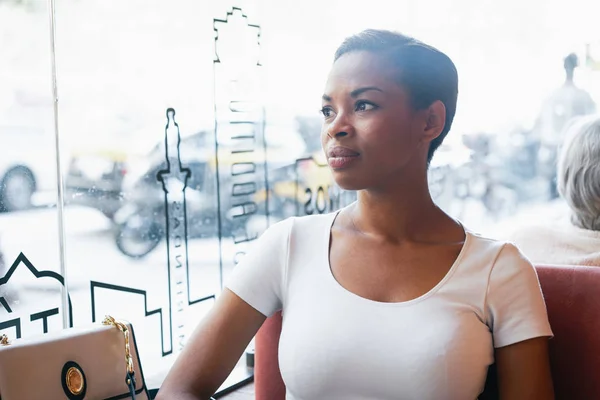 A beautiful african businesswoman with pulled back hair wearing white t-shirt is looking aside while sitting in a coffee shop
