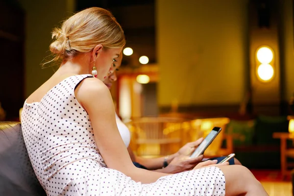 A side view photo of two female friends using electronic devices while sitting in a modern hotel lobby