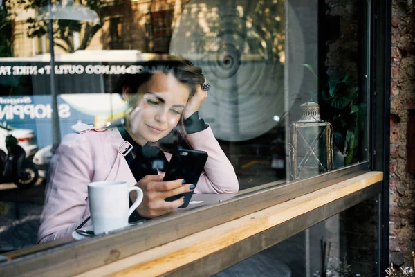 beautiful young woman writes a text message on a smartphone while sitting near the panoramic window in the cozy coffee house