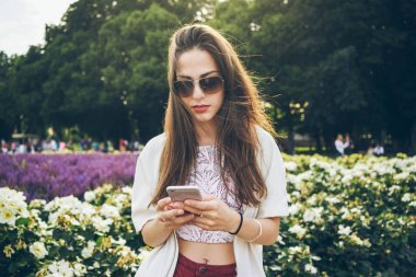 Güzel genç kadın şehir Garden'da dururken Smartphone kullanarak güneş gözlüğü takıyor