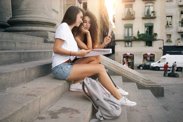 Young women sitting with a paper map and a modern smartphone and comparing the information from the different sources. Two female tourists are planning journey by using a map and a mobile phone