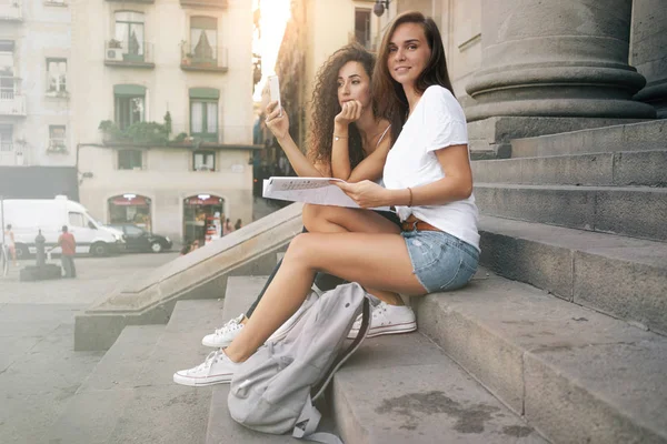 Two young women taking pictures while sitting outdoors on the stairs with backpacks and map.