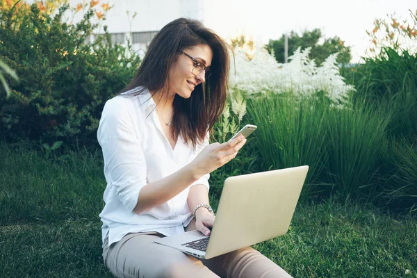 Young female student is looking at the screen of a smartphone while sitting on a grass with laptop on her knees