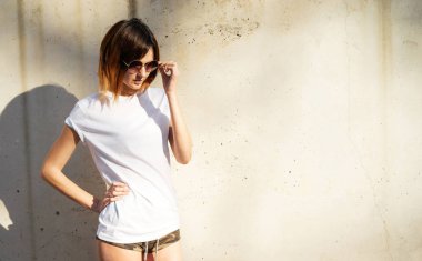 Beyaz boş t-shirt ve arka plan beton duvara karşı poz güneş gözlüğü giyen düzgün genç kadın