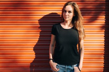 Uzun kahverengi saçlı bir turuncu duvar arka planda bir sokakta bekleyen bir boş siyah t-shirt giyen güzel genç kadın