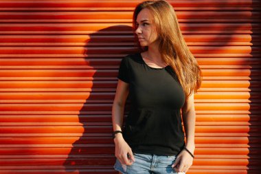 Uzun kahverengi saçlı bir turuncu duvar arka planda bir sokakta bekleyen bir boş siyah t-shirt giyen güzel genç kadın