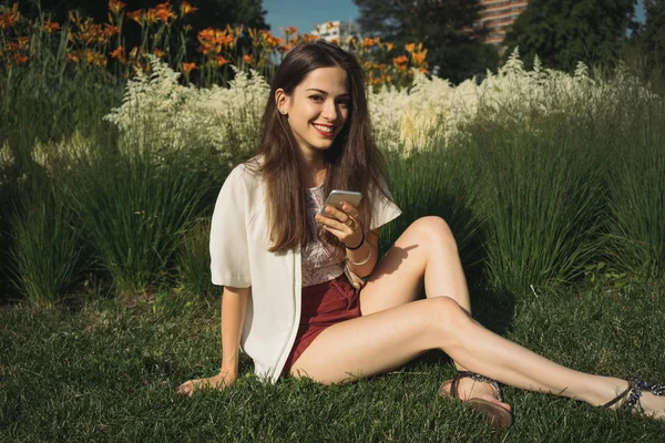 シティ ガーデンで座りながらスマート フォンを使用してサングラスをかけて美しい若い女性 — ストック写真