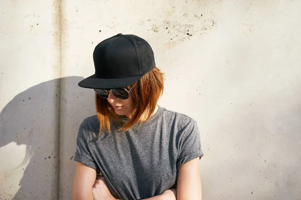 美丽的年轻女子穿着灰色的 T恤衫和黑色的帽子摆在一个混凝土墙的背景 — 图库照片