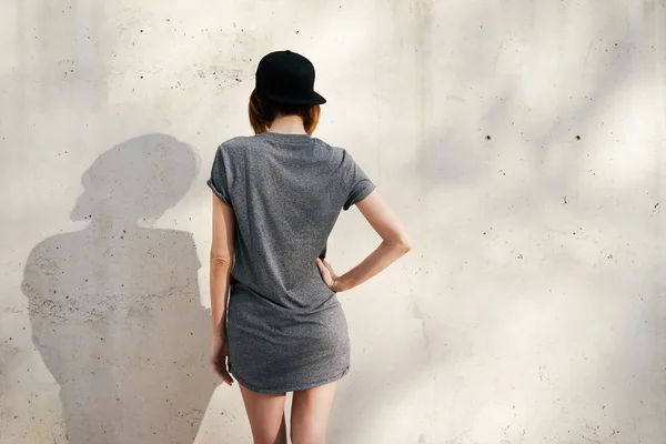 美丽的年轻女子穿着灰色的空白 T恤衫和黑色帽子摆在混凝土墙的背景上 — 图库照片