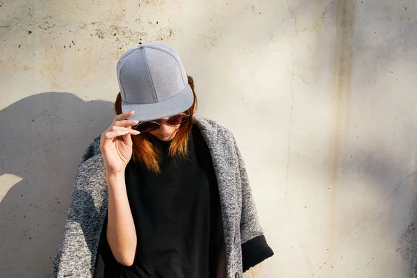 Young woman  wearing a grey blank cap and sunglasses posing against concrete wall background