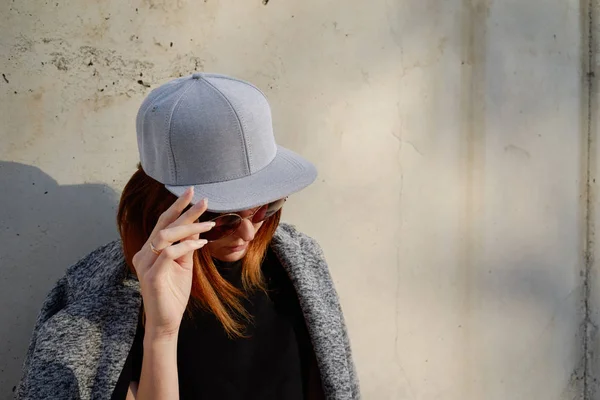 戴灰色空白帽和太阳镜的年轻女子反对混凝土墙背景 — 图库照片