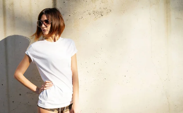 Shapely young woman wearing white blank t-shirt and sunglasses posing against a background of a concrete wall