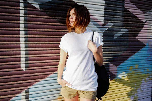 young attractive woman with a backpack wearing white t-shirt standing on a graffiti wall background